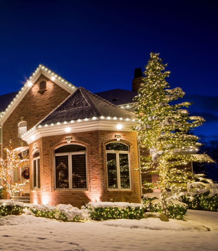 Warm white holiday lights along a brick house roof, hedges, and a snow-covered evergreen